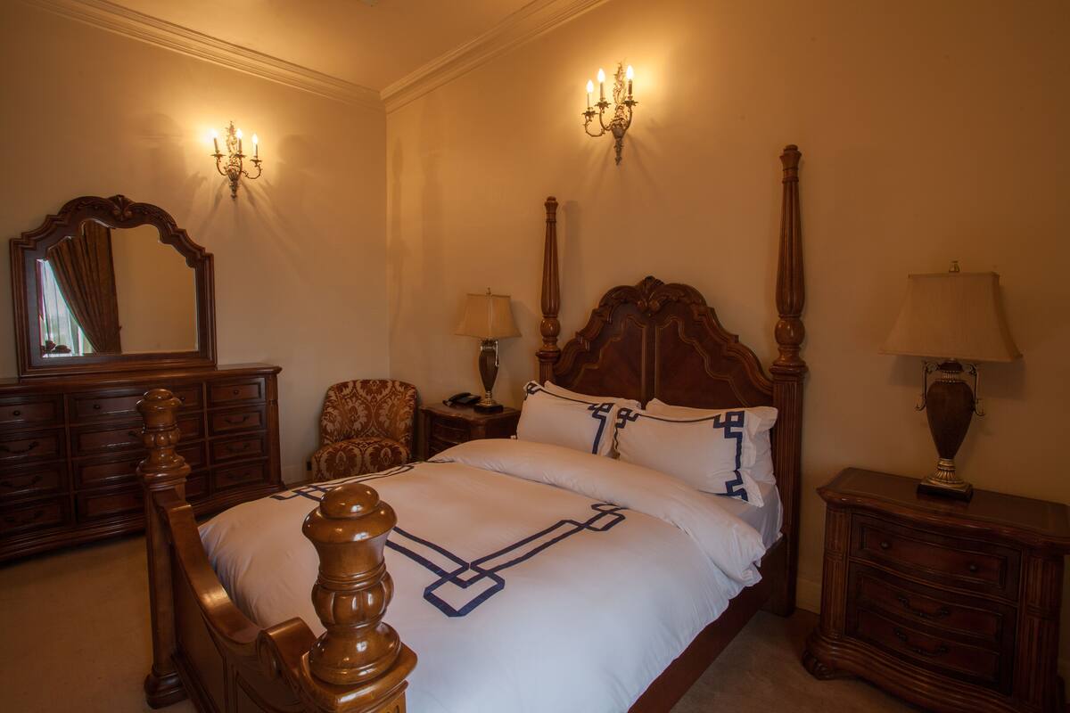 A bedroom with a large bed, wooden headboard, two bedside tables with lamps, a chair, a dresser, and a mirror on the wall.