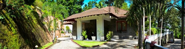 Un bâtiment de style traditionnel, doté d’un toit en tuiles, entouré d’une végétation luxuriante et accessible par un chemin pavé menant à l’entrée.