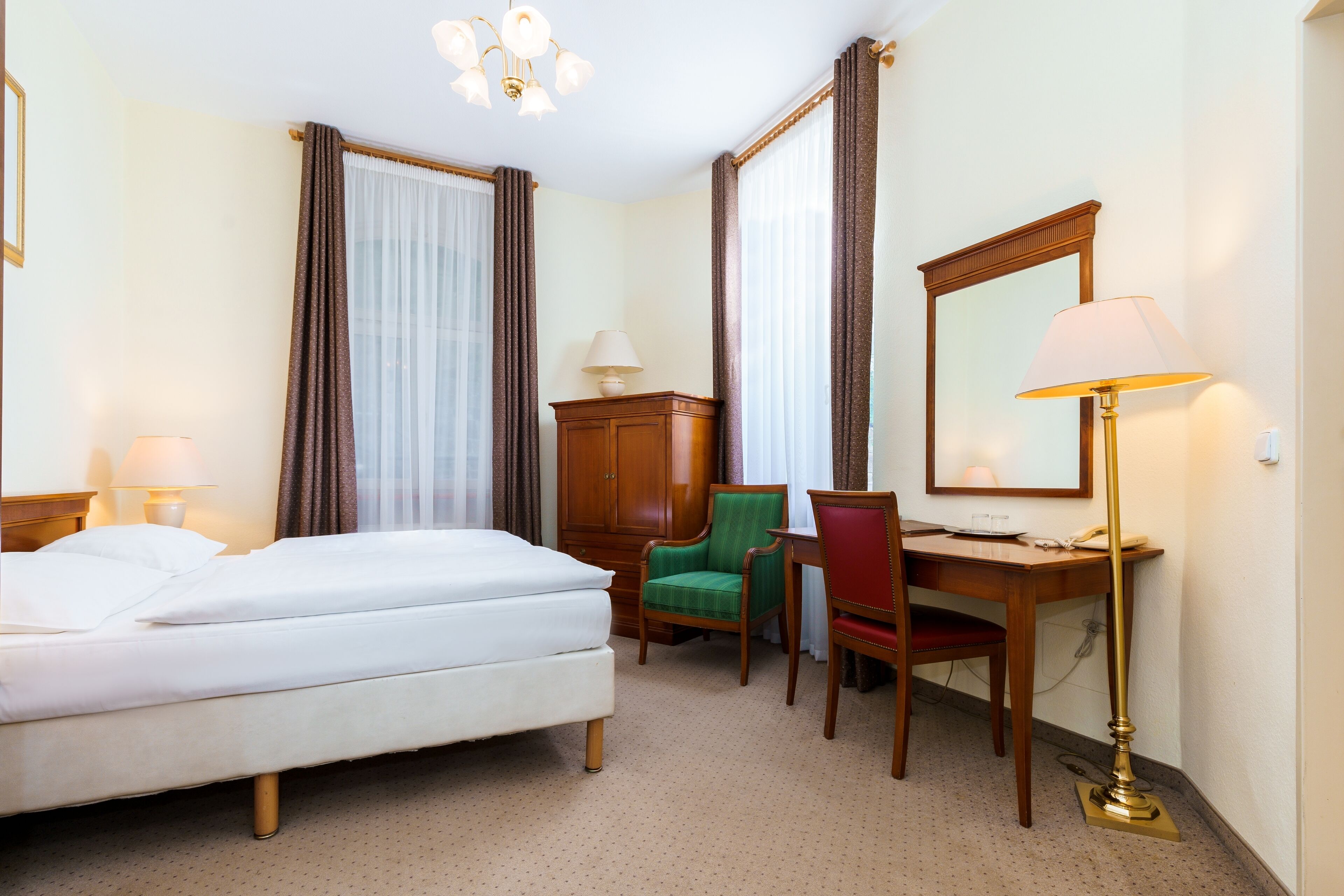 A hotel room with a bed, a desk, a chair, a lamp, a mirror, and a wardrobe.