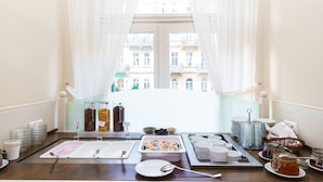 A breakfast buffet setup with various food items and utensils, a view of a building through sheer curtains, and a window with a chandelier. - Hotel Anglický Dvůr (Karlovy Vary)