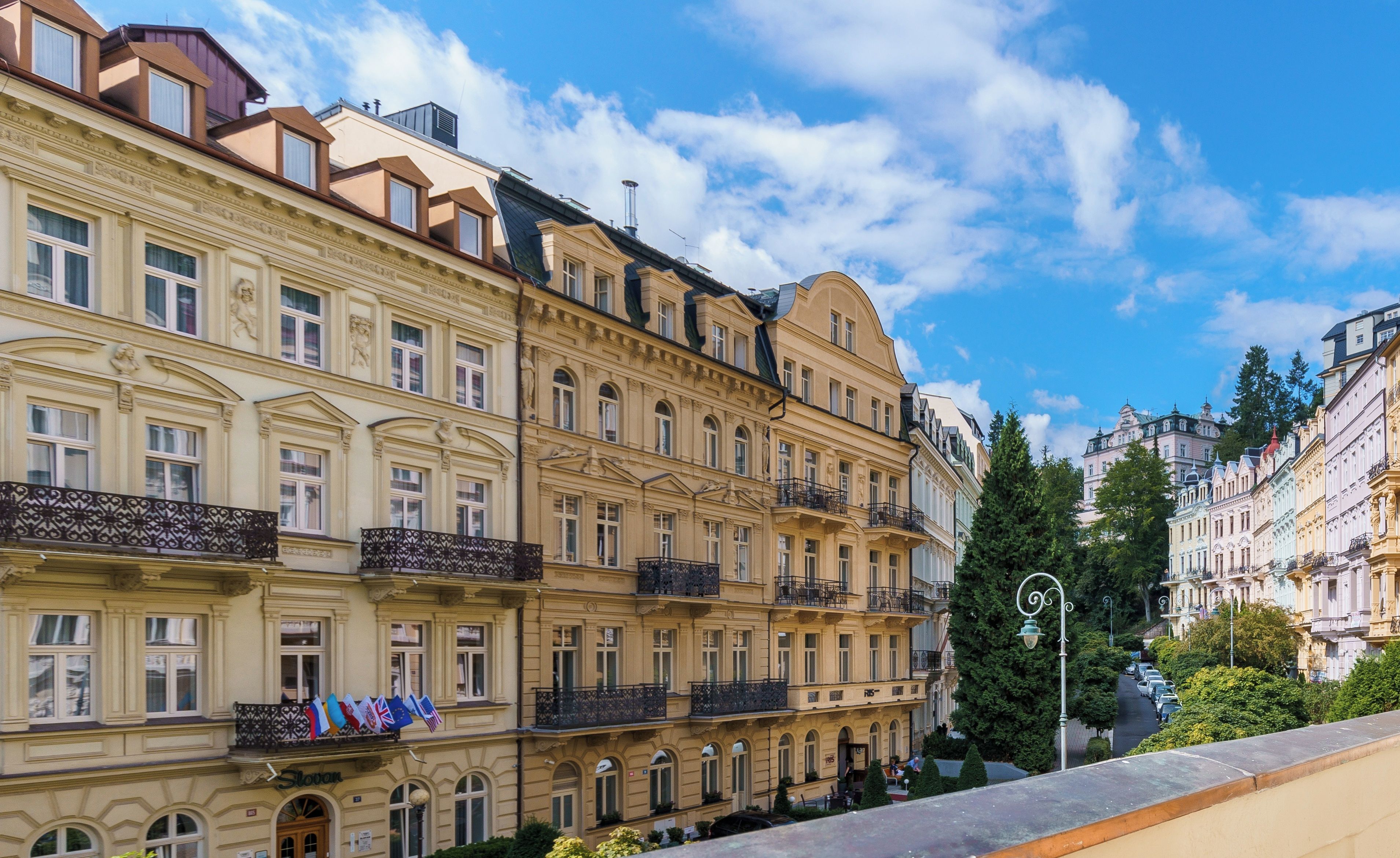 Fila de edifícios altos com sacadas, uma rua em primeiro plano e um céu azul claro.