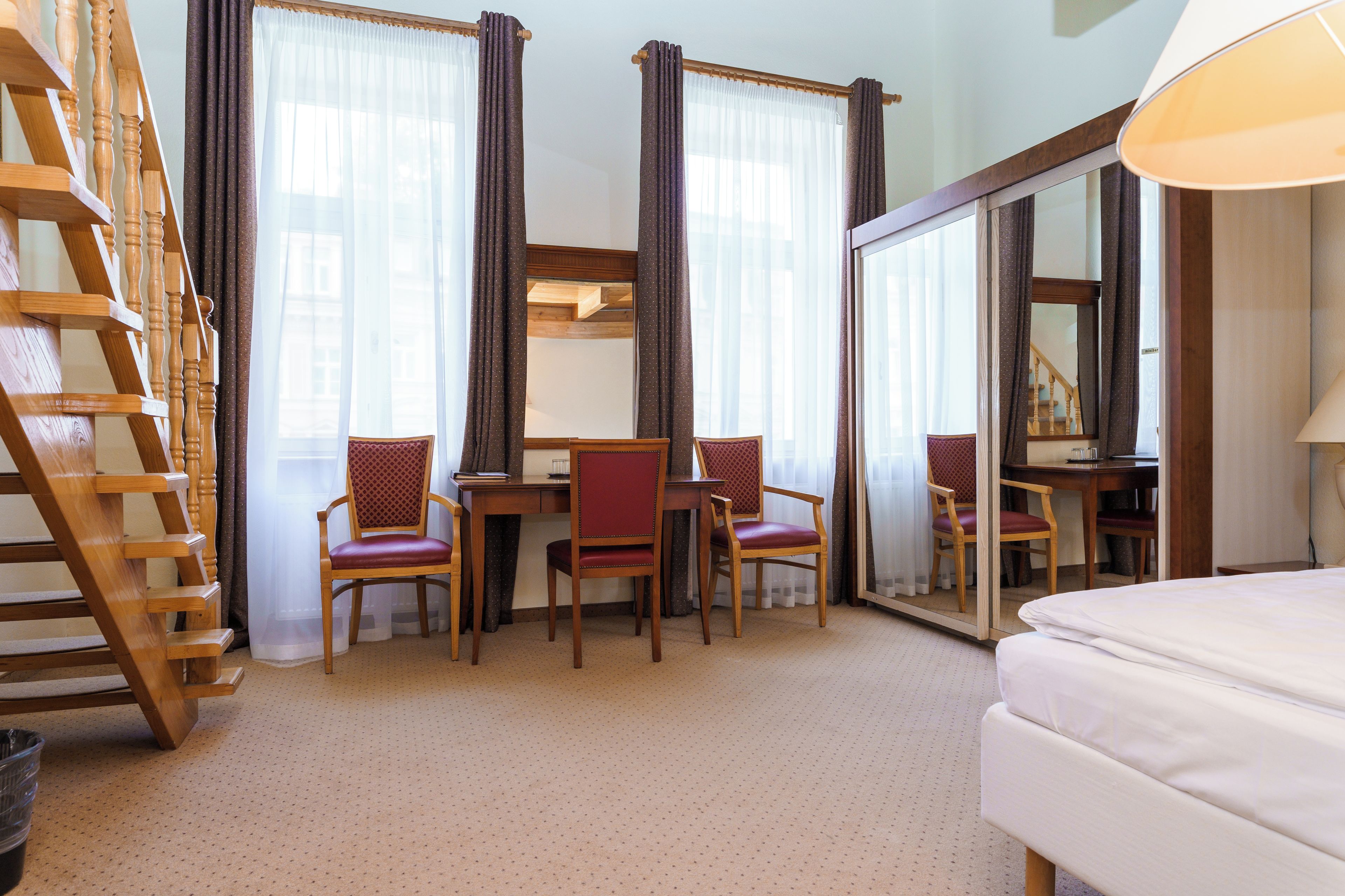 A hotel room with a wooden staircase, a desk with two chairs, a bed, and large windows with curtains.