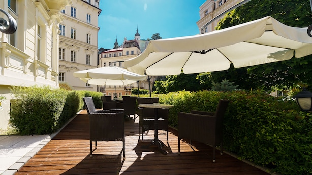 An outdoor patio with wicker furniture and white umbrellas, surrounded by greenery and historic buildings. - Hotel Anglický Dvůr (Karlovy Vary)