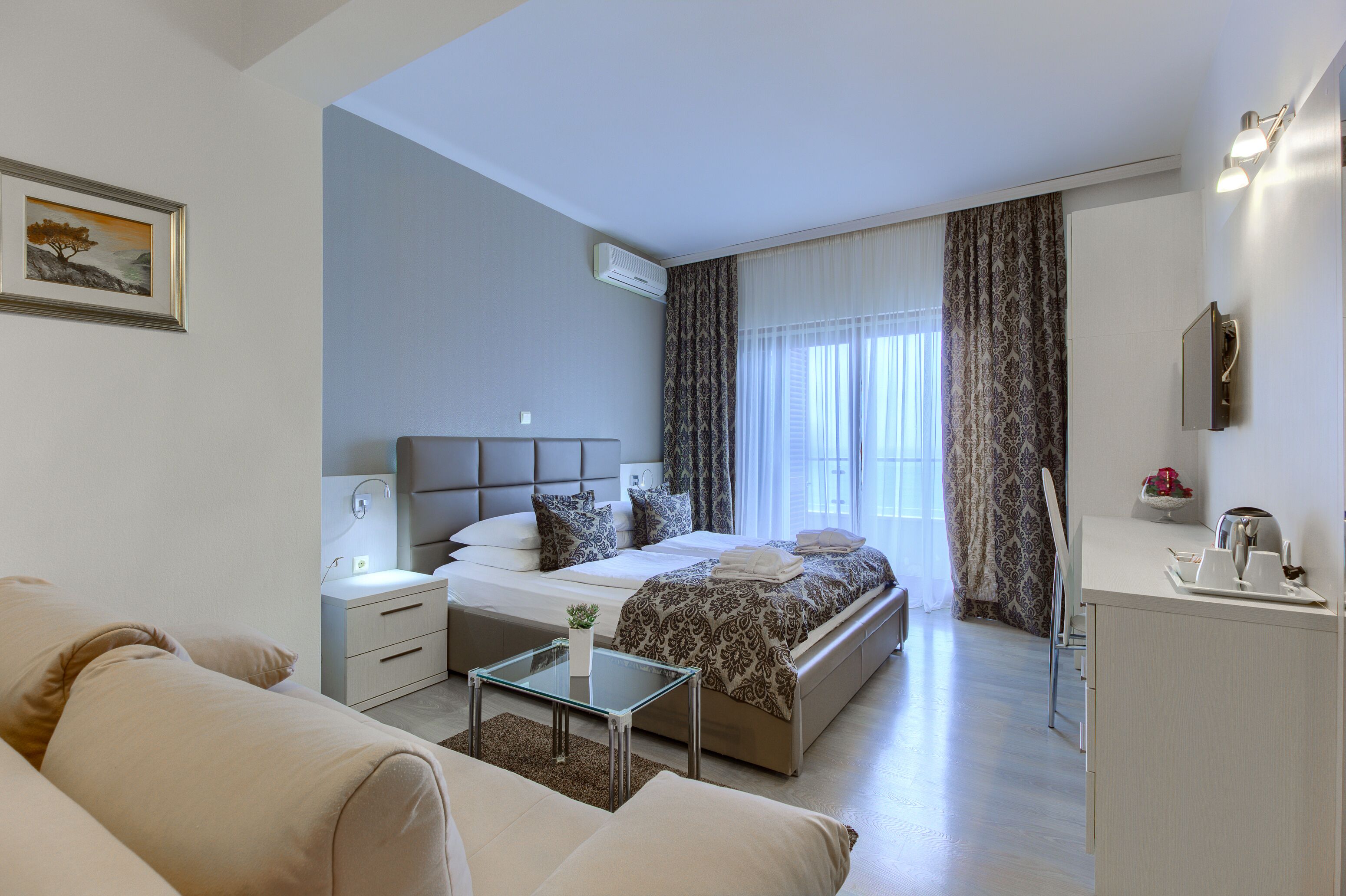 A modern hotel room with a large bed, a bedside table, a small table with a plant, and a desk with a lamp.