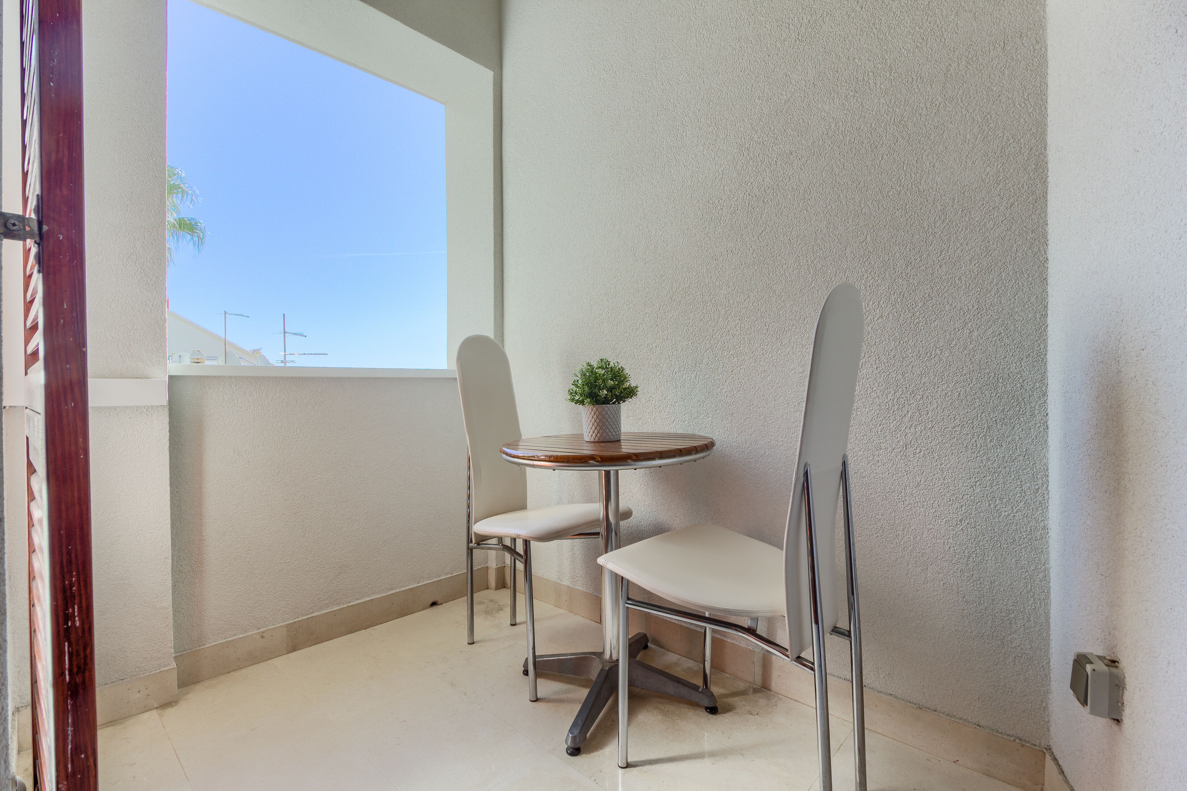 A small balcony with a white chair and a table, a potted plant, and a view of the sky.