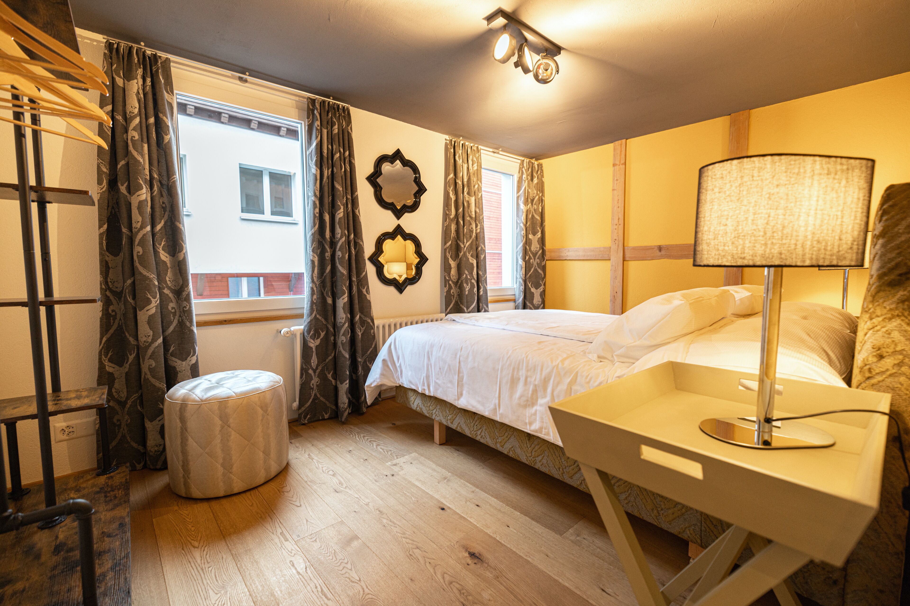 A hotel room with a bed, bedside table with a lamp, a small ottoman, a window with curtains, and a wall-mounted mirror.