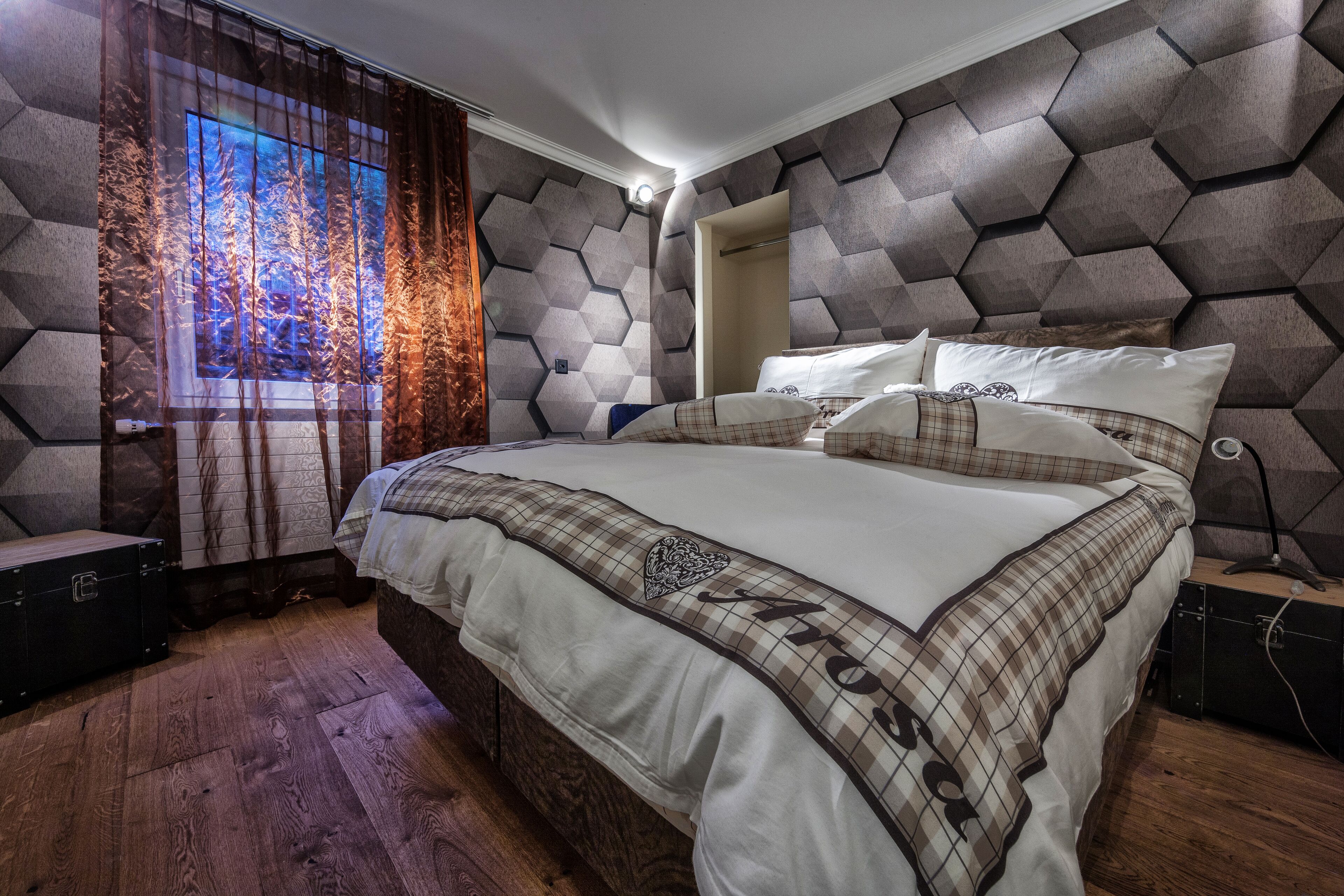 Een moderne slaapkamer met een hoofdeinde met een geometrisch patroon, een groot bed met ruitbeddengoed en uitzicht op een bos door een raam met dunne gordijnen.