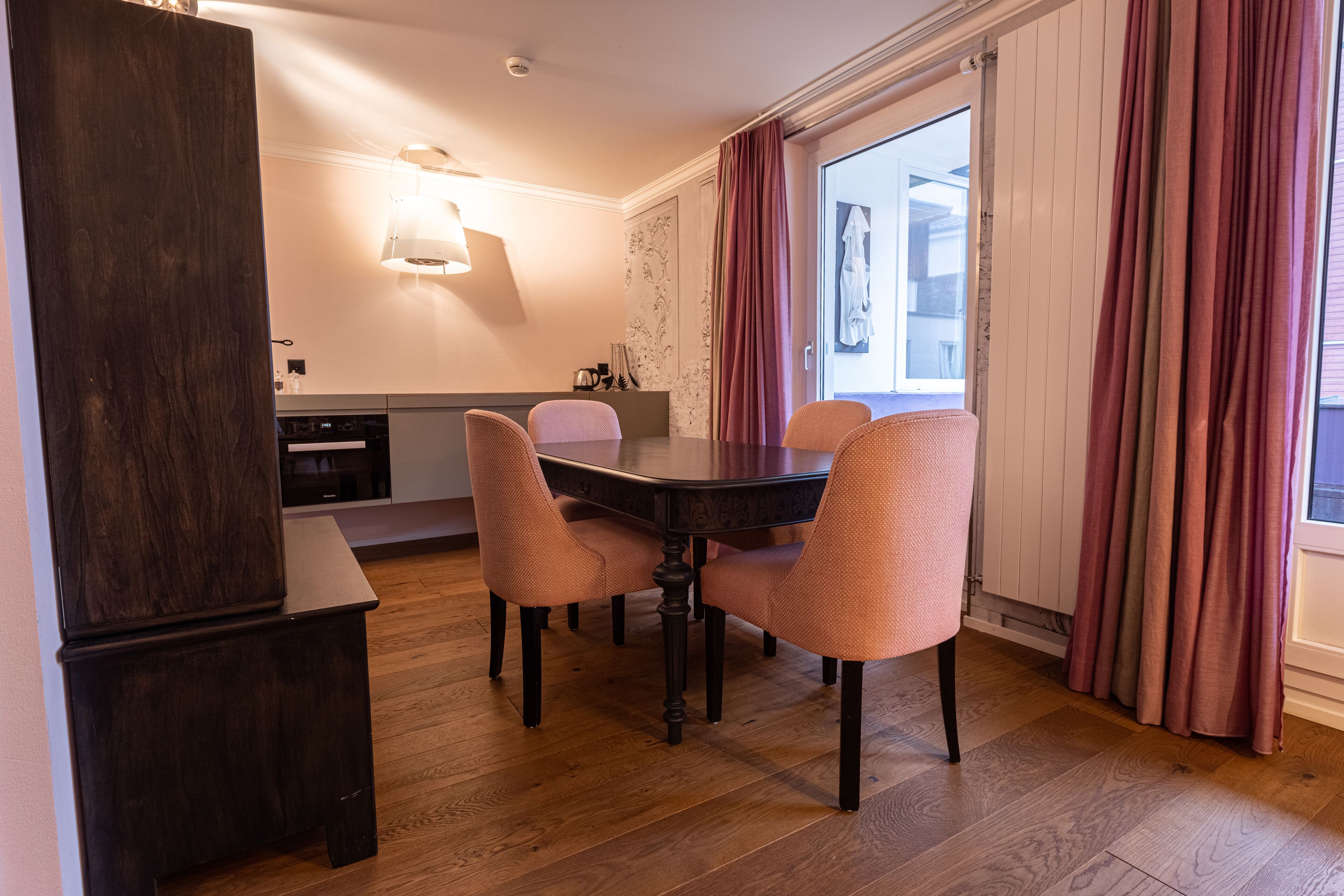Un comedor con una mesa de madera oscura y cuatro sillas acolchadas. Una lámpara moderna cuelga sobre la mesa, y se ve una ventana con cortinas a la derecha.