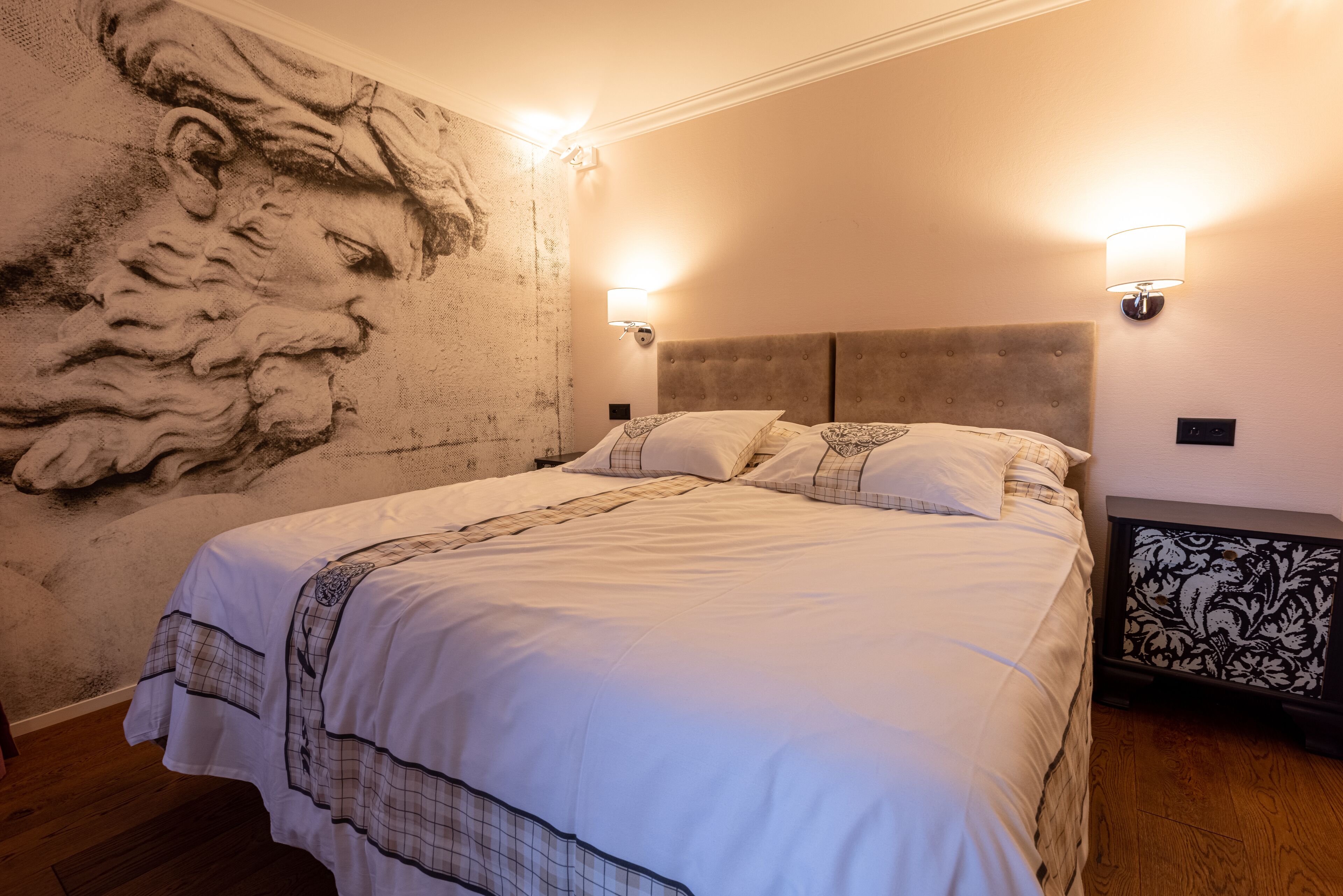 A bedroom with a large bed, two wall-mounted lights, a headboard, and a decorative wall mural.