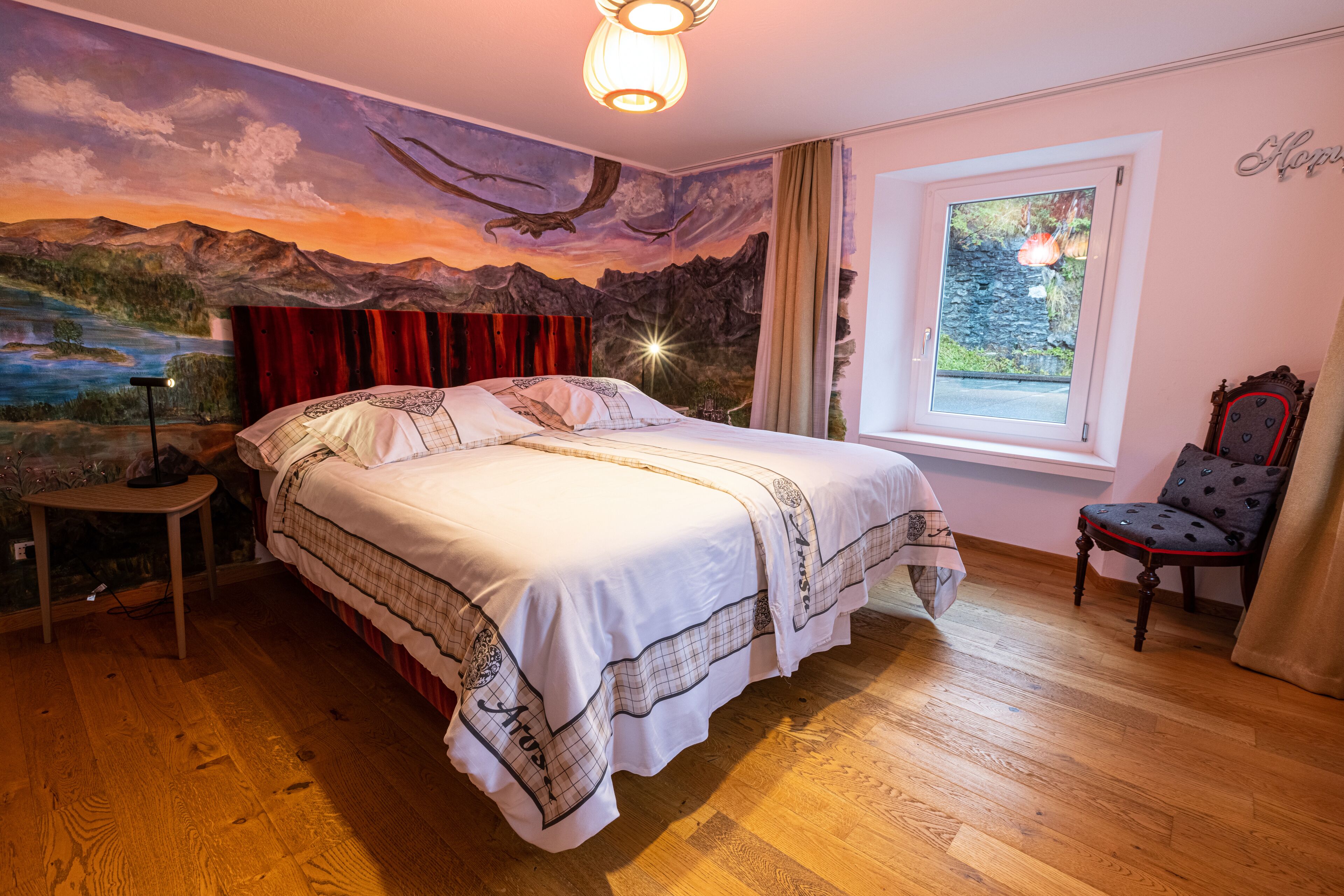 A bedroom with a mural of a sunset landscape, a bed with a white and patterned bedspread, a wooden floor, a small table with a lamp, and a window with a view of greenery.