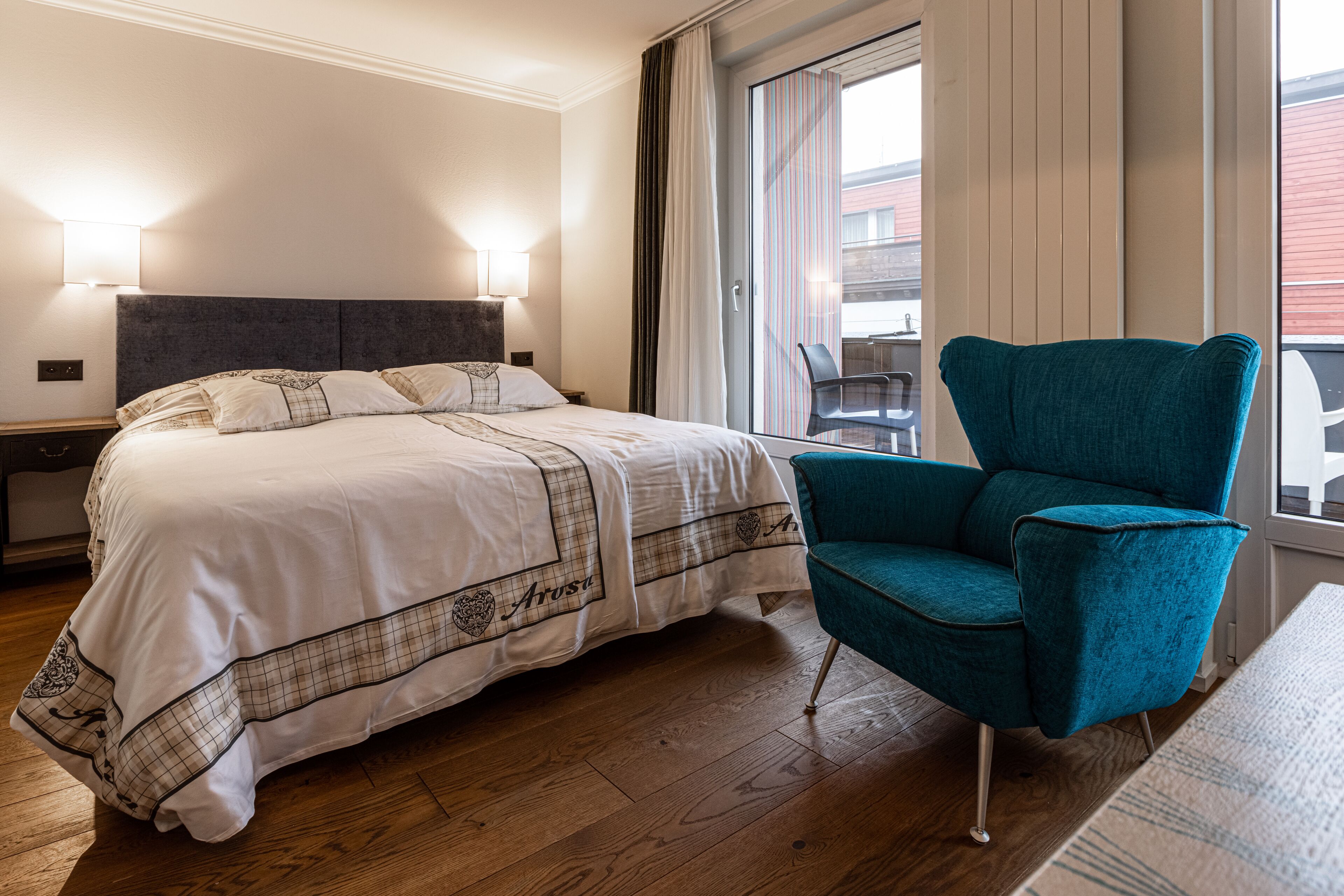 A hotel room with a large bed, a teal armchair, a nightstand, and a window with curtains.