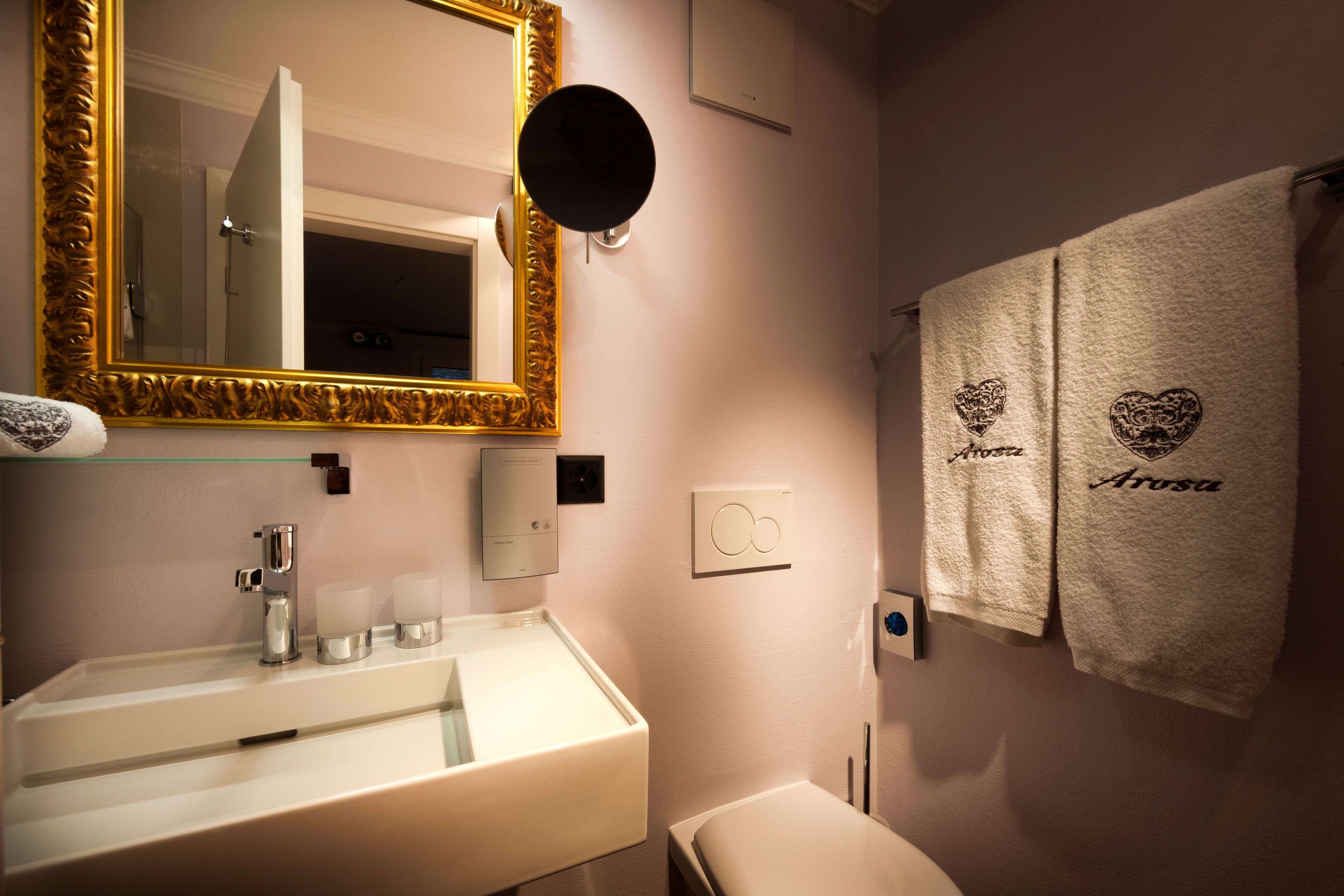 A bathroom with a white sink, a gold-framed mirror, and two towels with 'Arosa' embroidered on them.