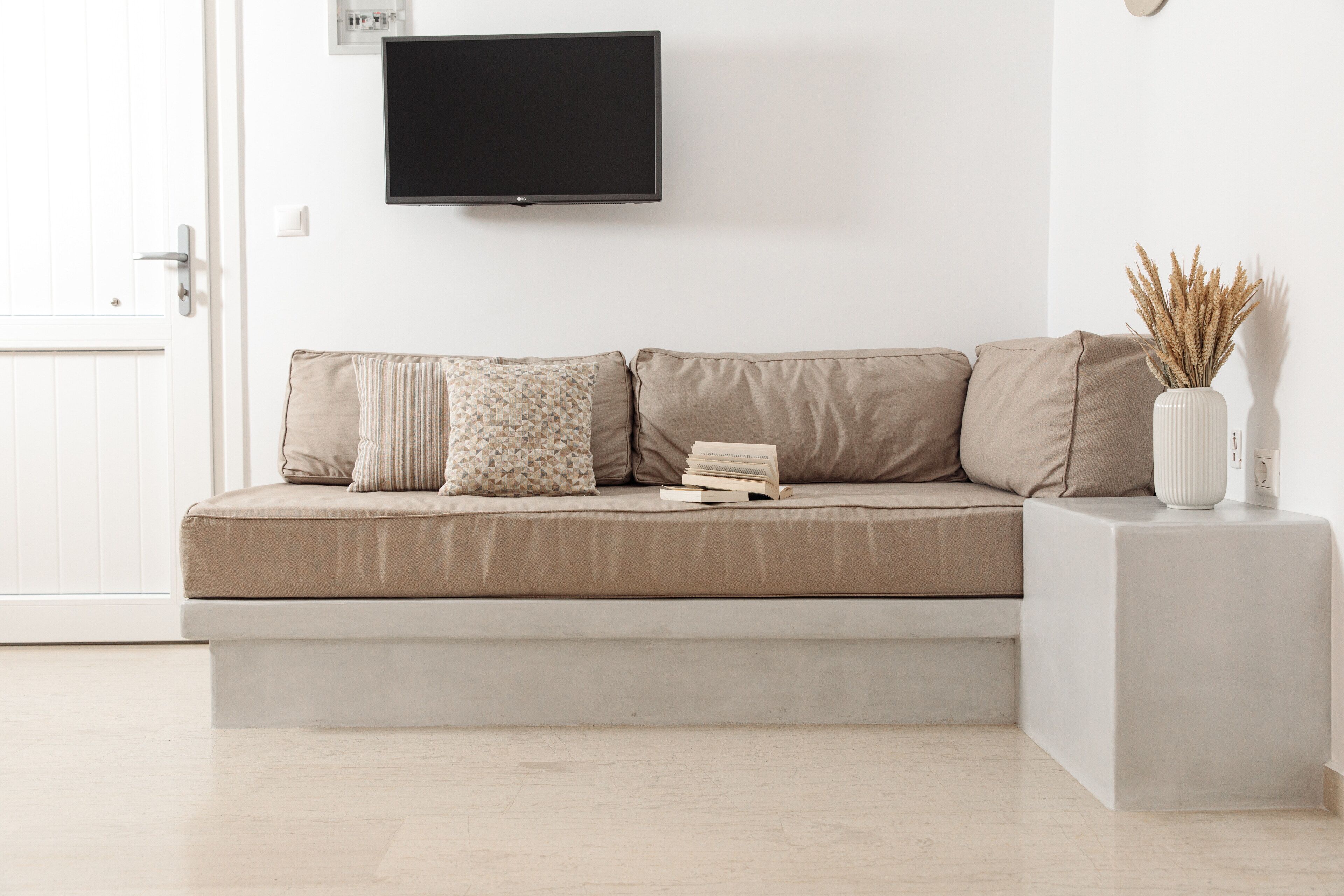 Ein schlichter Wohnraum mit einem beigefarbenen Sofa, einem Wand-Fernseher und einer weißen Vase mit Trockenpflanzen auf einem Beistelltisch.