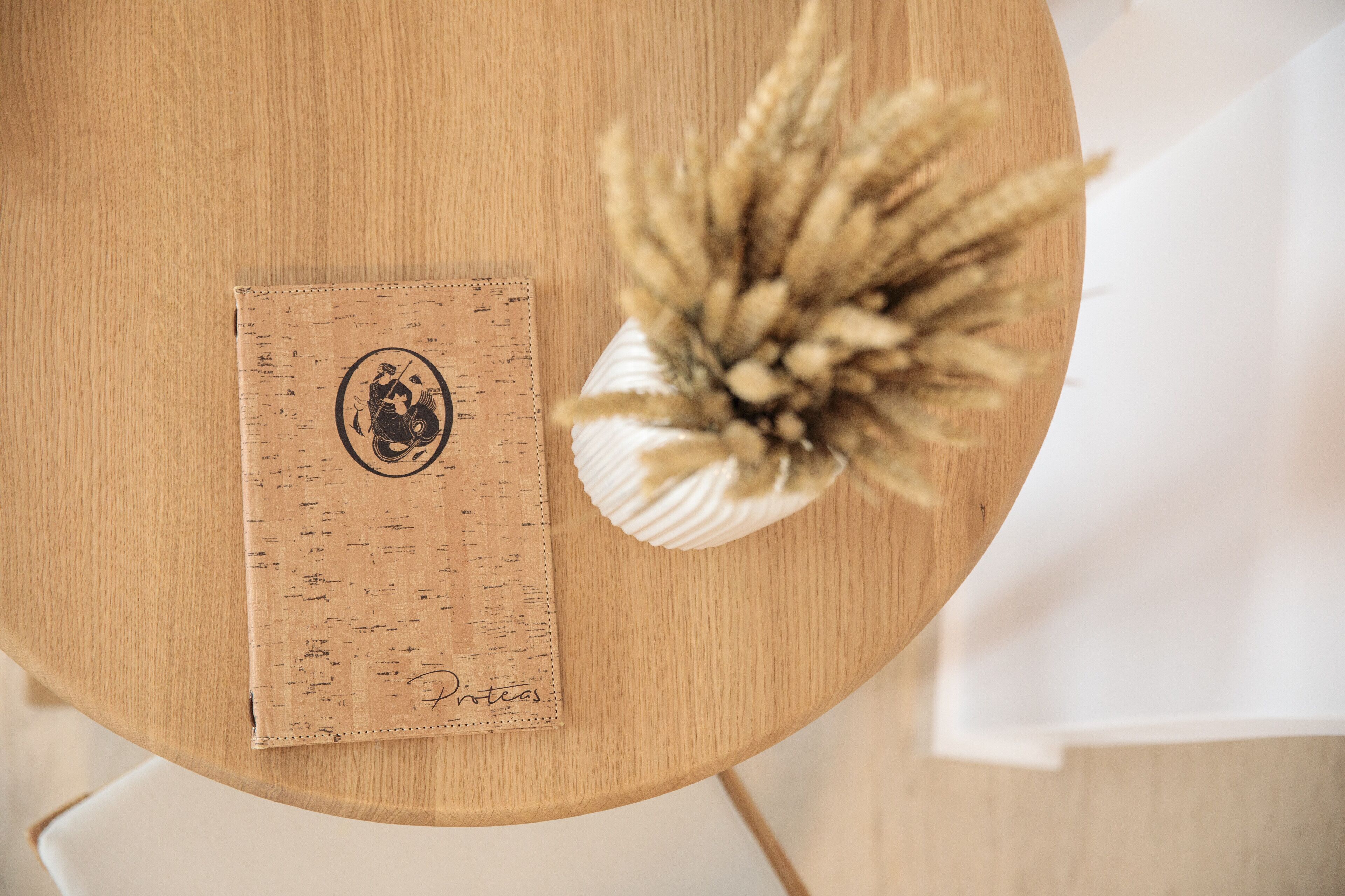 A round wooden table with a corkboard, a decorative vase with dried wheat, and a piece of paper with a signature.
