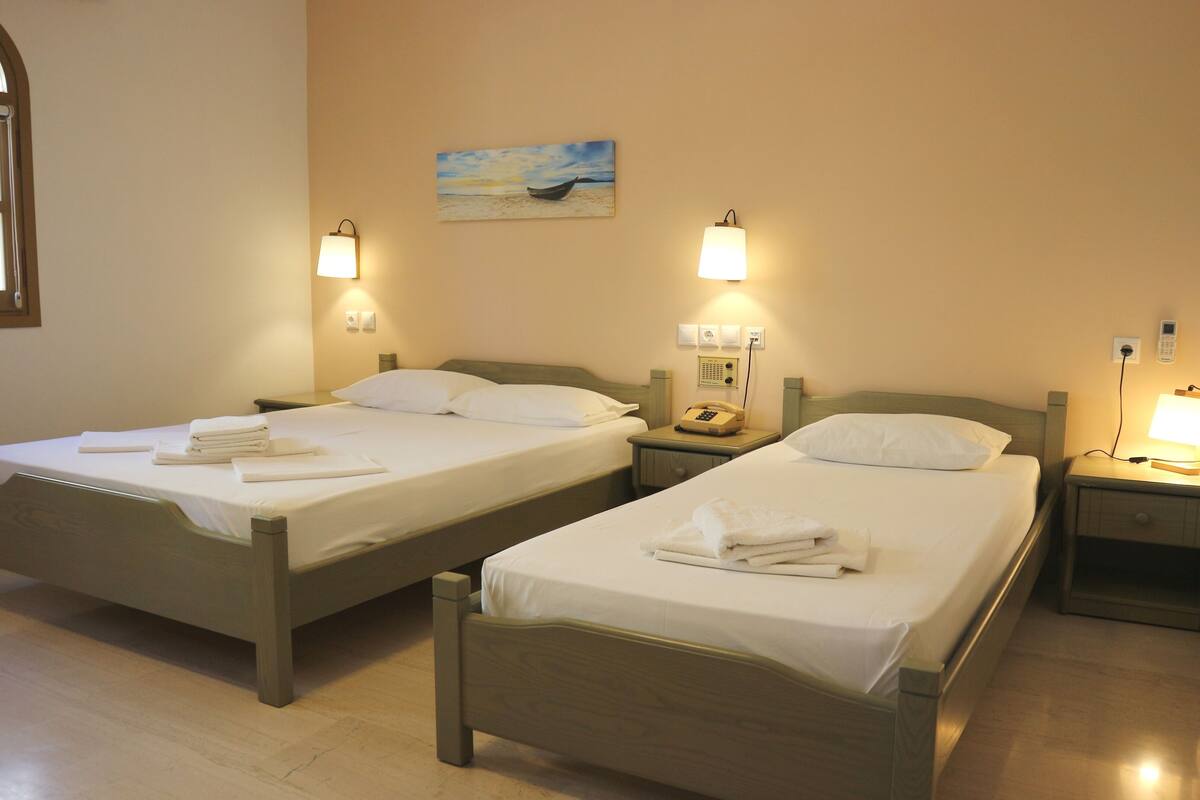Two single beds with wooden frames, white linens, and towels, bedside tables with lamps, a painting of a boat on the wall, and a window with a view of the sea.