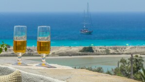 To glas øl på en stenkant med udsigt til en strand og en sejlbåd i det fjerne.