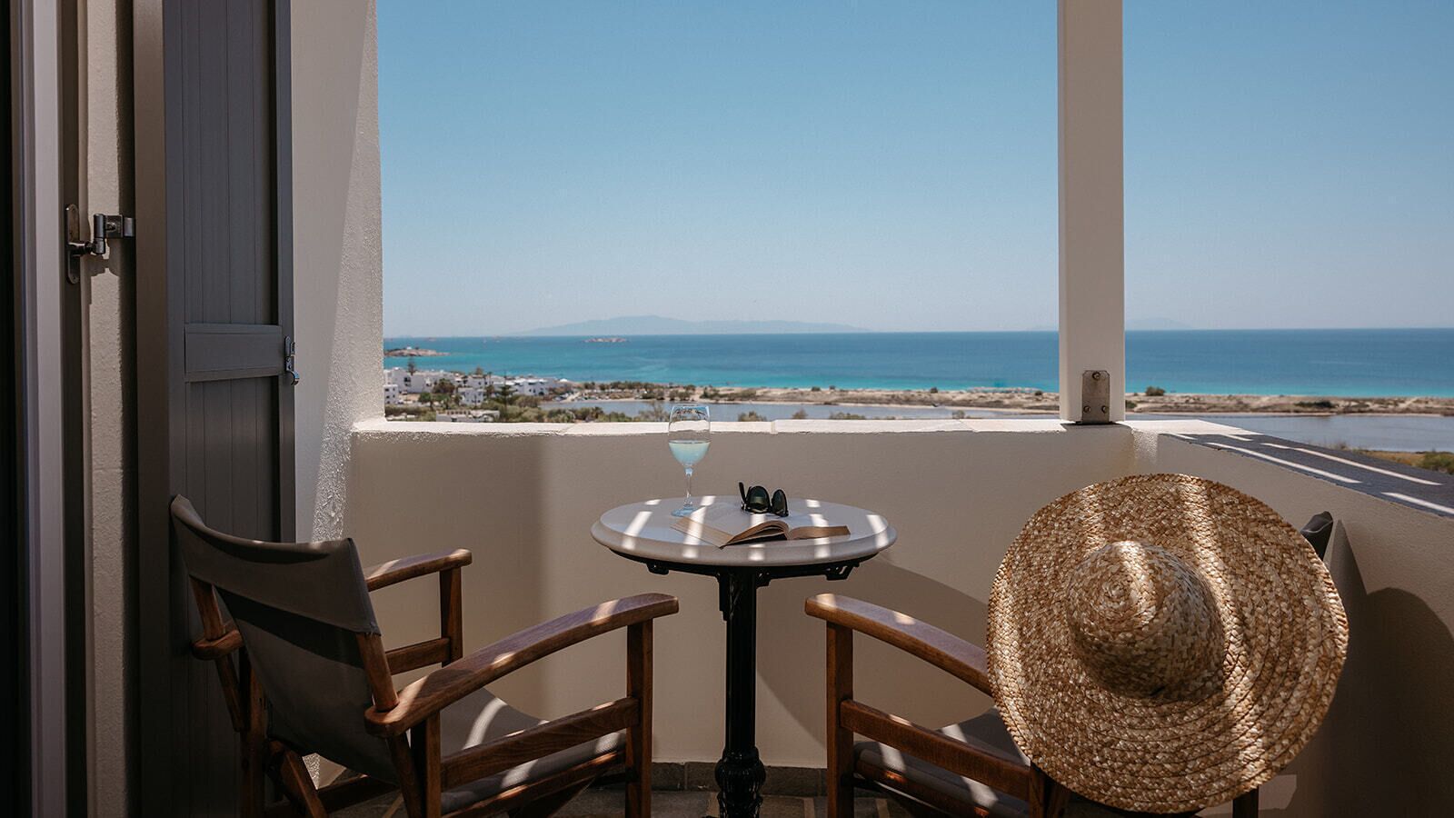 A balcony with two wooden chairs, a round table, a straw hat, and a view of the sea and distant land.