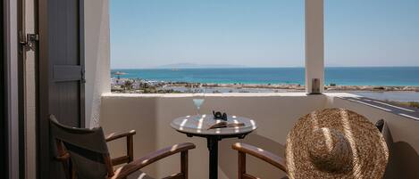 A balcony with two wooden chairs, a round table, a straw hat, and a view of the sea and distant land.