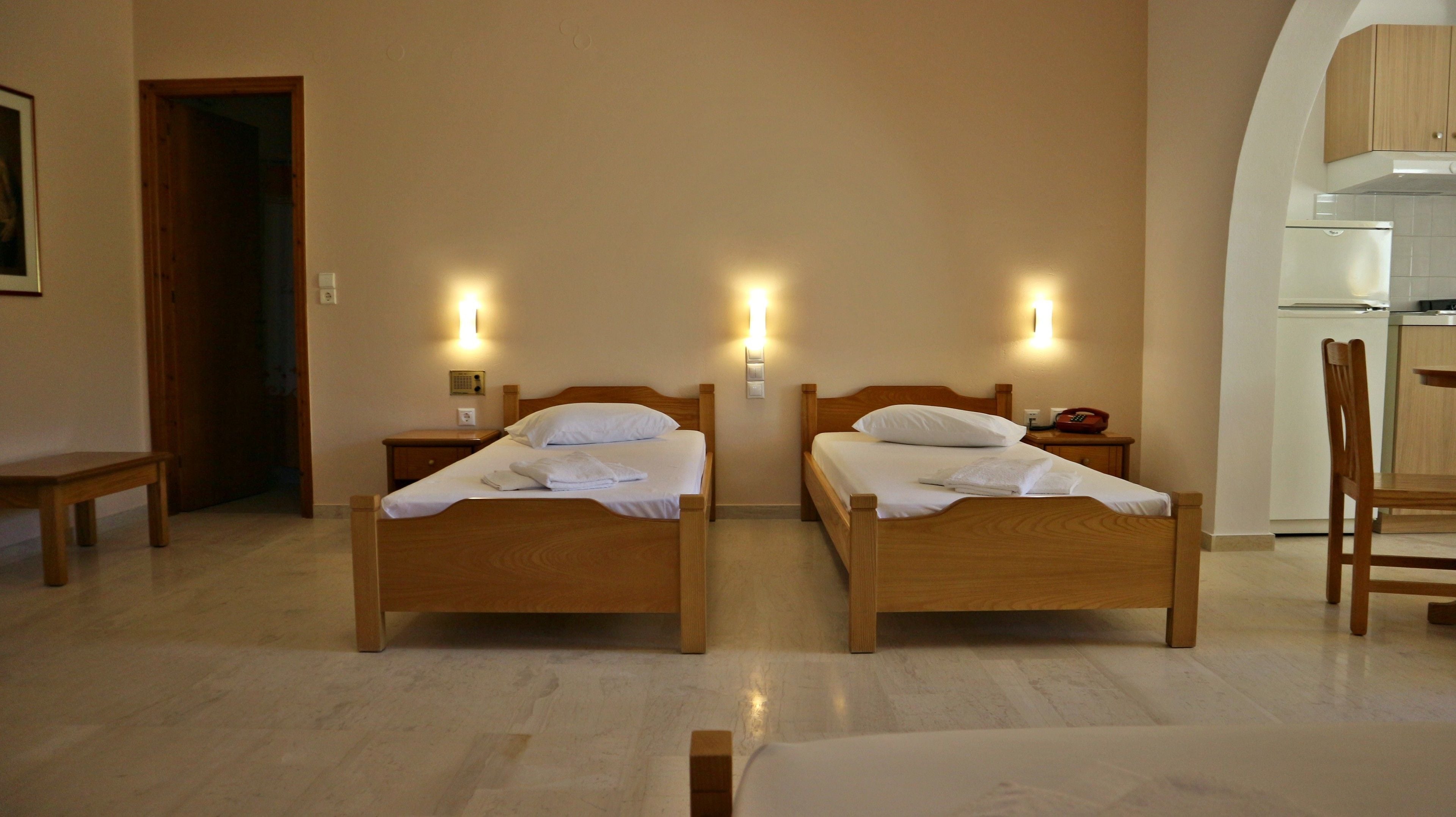 A hotel room with two wooden beds, a small table, a chair, and a kitchenette.