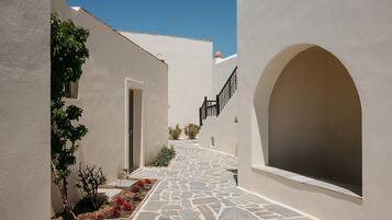 A narrow stone-paved alleyway between white buildings with arched doorways and a small garden area with red flowers.