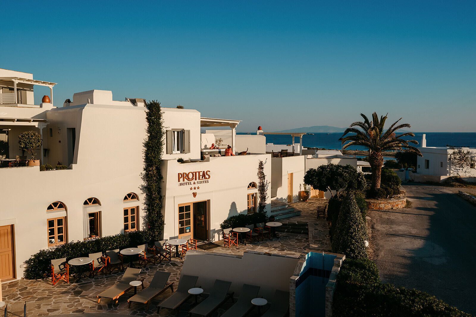 A hotel named 'PROTEAS HOTEL & SUITES' with outdoor seating and a view of the sea.