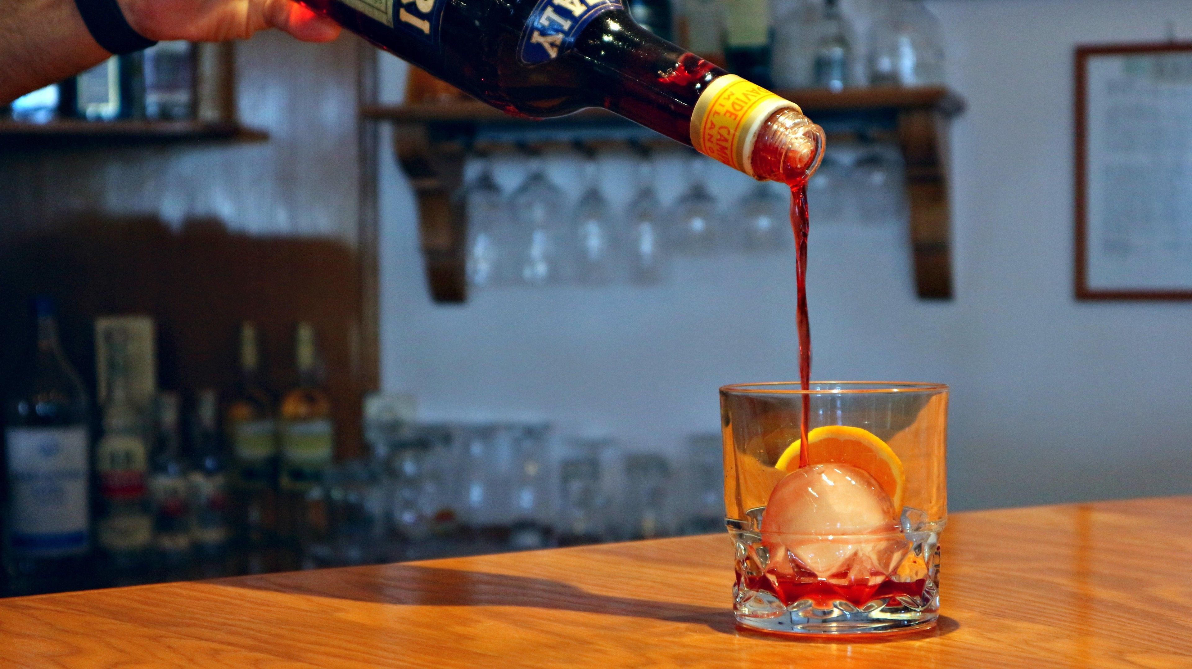 A person pouring a red liquid into a glass with an ice cube and an orange slice.