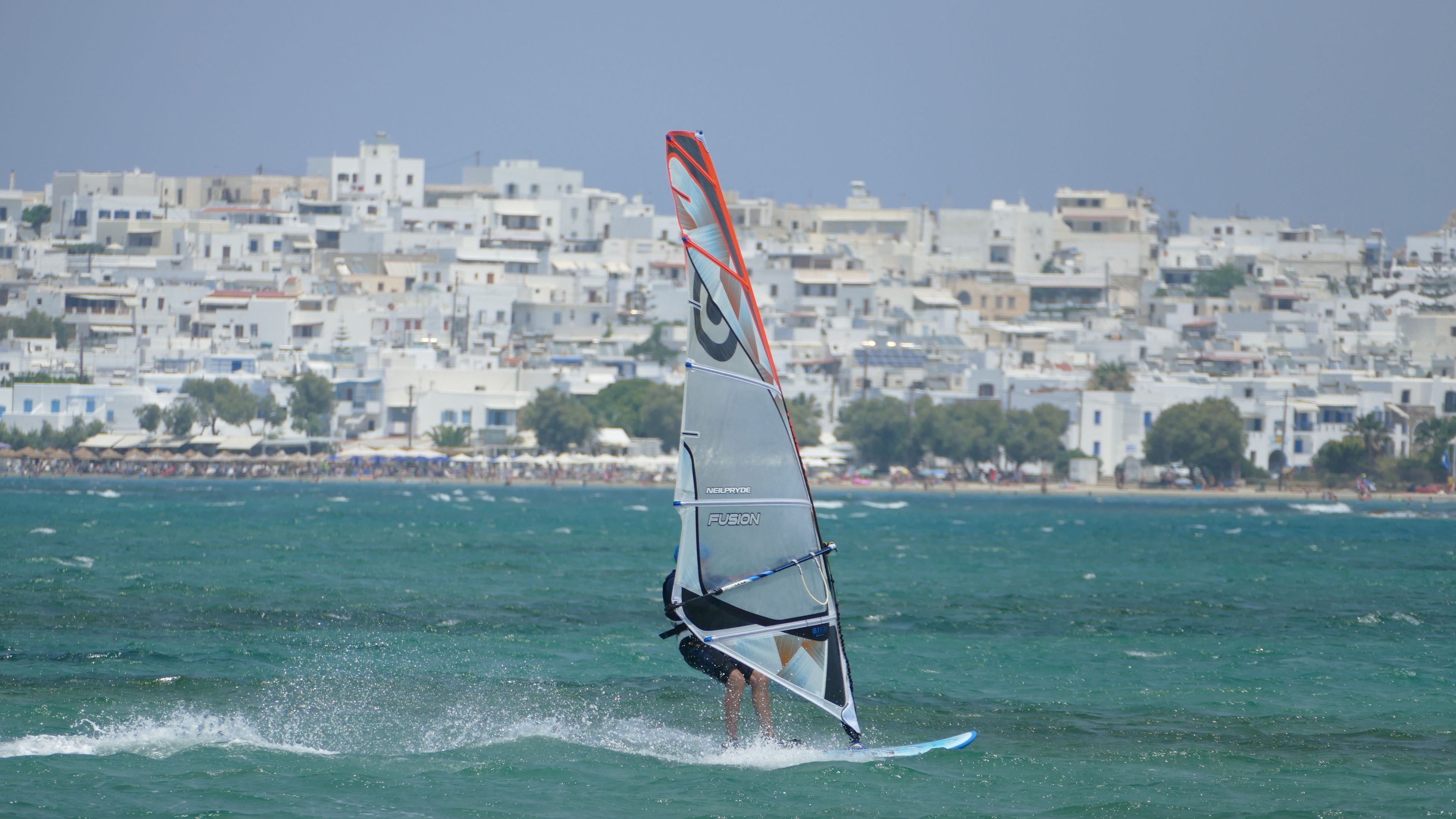 Une scène de planche à voile avec un voilier sur l’eau et une ville côtière en arrière-plan.