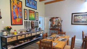 A dining area with a wooden table and chairs, a buffet counter with food items, and various artworks on the walls. - Destiny Inn Lodge (Polokwane)