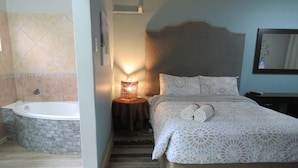 A bedroom with a bed, a nightstand, a lamp, a wall-mounted mirror, and a bathroom with a bathtub and a towel rack. - Destiny Inn Lodge (Polokwane)