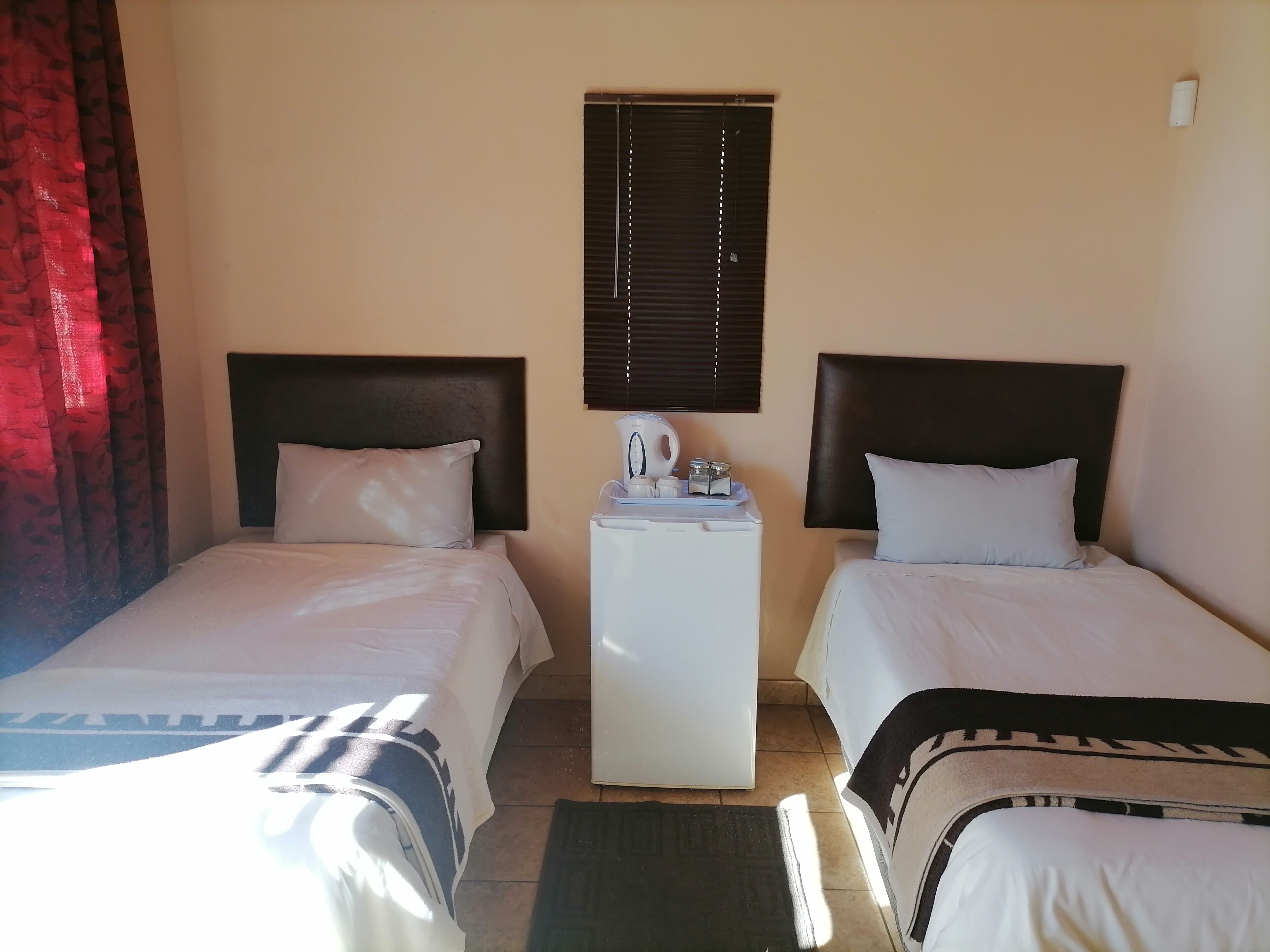 A hotel room with two beds, a small table with a kettle, and a window with blinds.