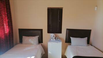 A hotel room with two beds, a small table with a kettle, and a window with blinds.