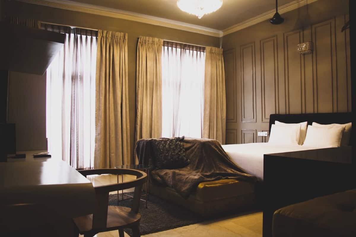 Une chambre d’hôtel avec un lit, un bureau et une chaise. Les fenêtres sont dotées de rideaux, et un lustre est suspendu au plafond.