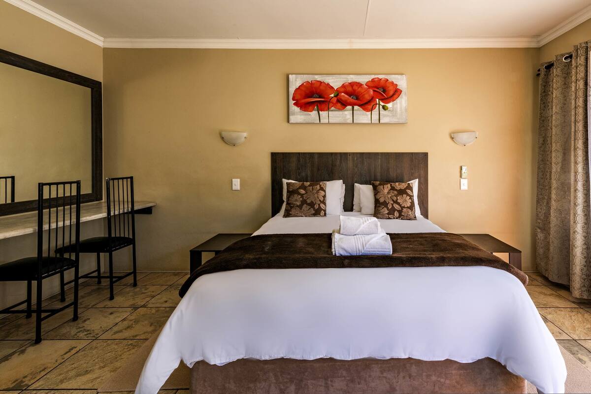 Ein Hotelzimmer mit einem Bett, zwei Stühlen, einem Bild mit roten Blumen und einem Fenster mit Vorhängen.