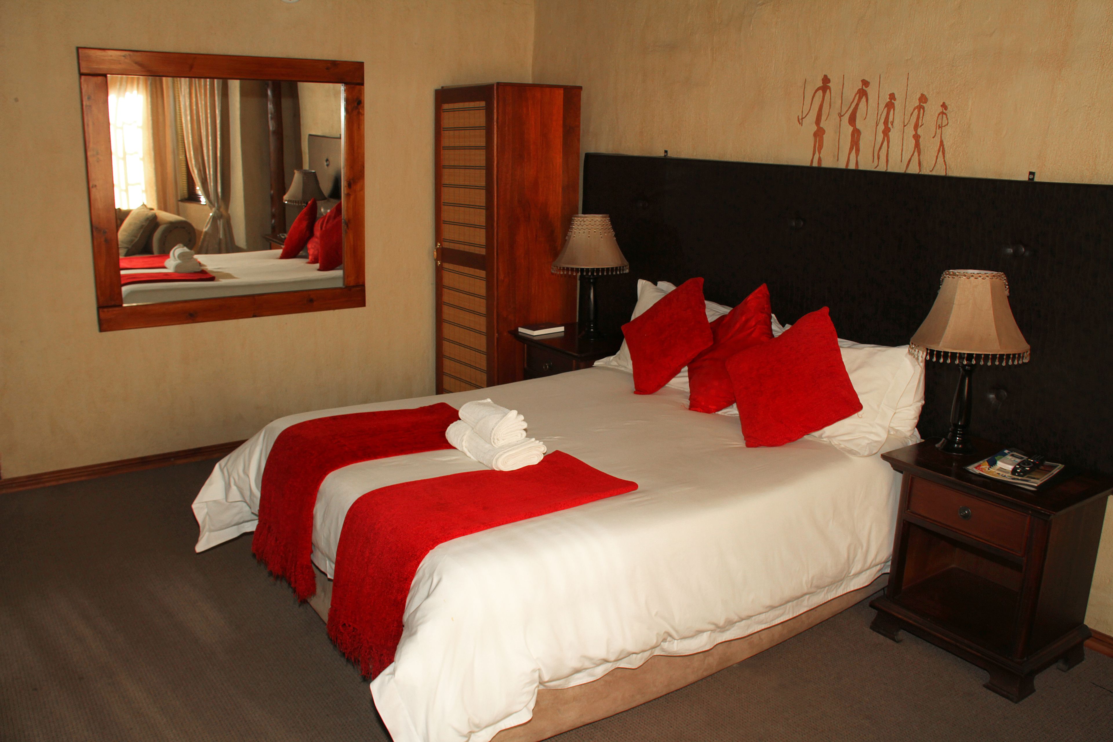 Een netjes opgemaakt bed met rode en witte beddengoed, een houten nachtkastje met een lamp en een spiegel aan de muur.