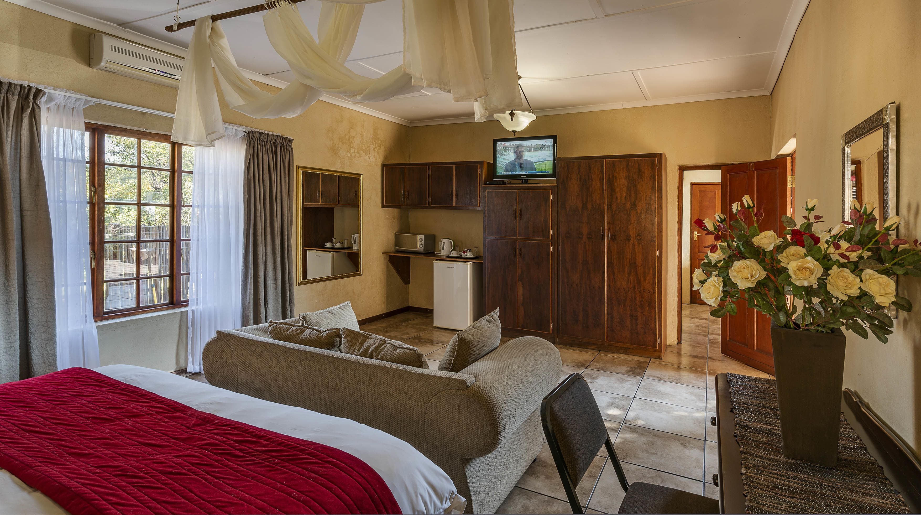 A bedroom with a bed, a red blanket, a sofa, a TV, a small kitchenette, and a door leading to another room.