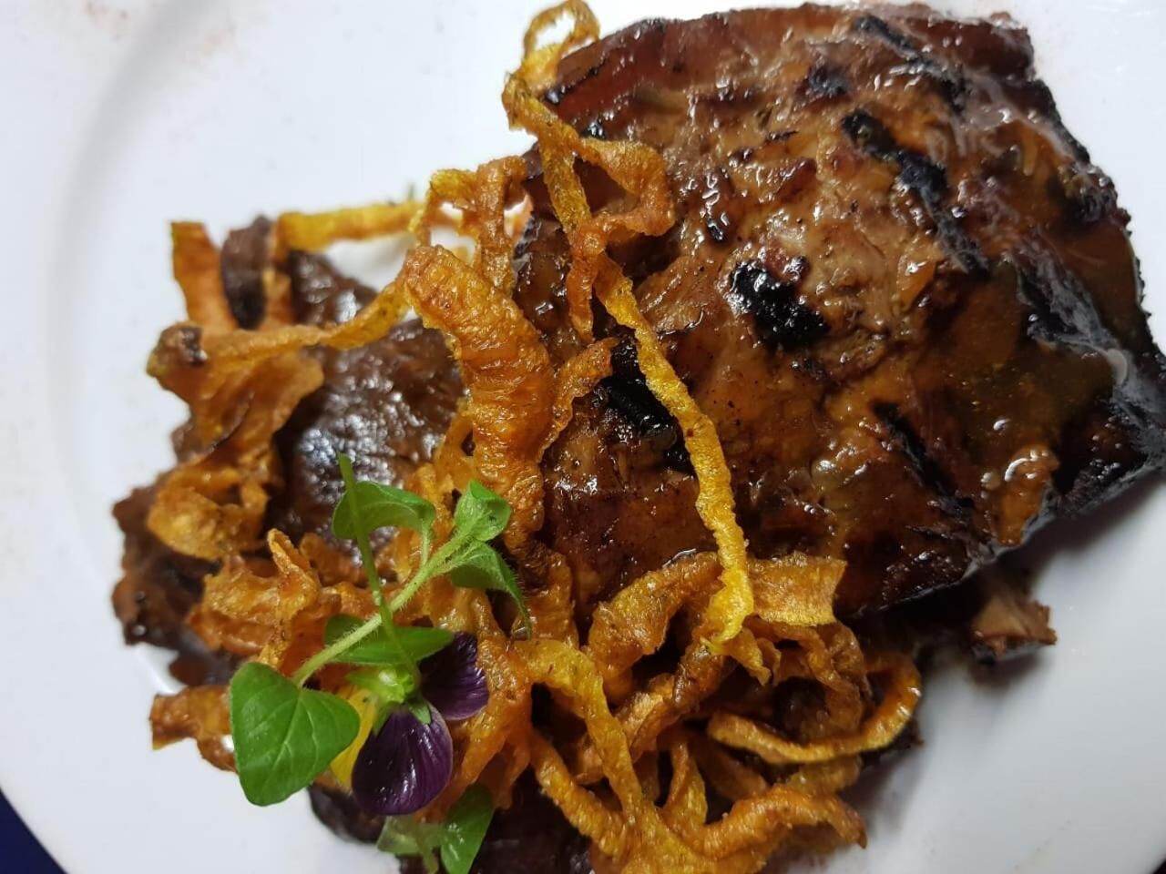 Un plato con carne a la parrilla cubierta de cebollas caramelizadas y adornada con una pequeña hierba verde.