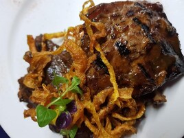 Un plato con carne a la parrilla cubierta de cebollas caramelizadas y adornada con una pequeña hierba verde.