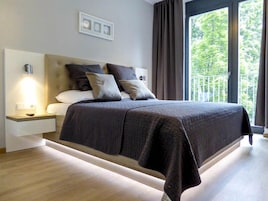 A modern bedroom with a large bed, a nightstand with a lamp, and a view of greenery through the window.