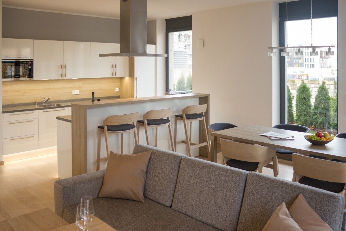 Une cuisine moderne avec des meubles blancs, un îlot central doté de chaises, une table à manger et une vue sur les arbres à travers de grandes fenêtres.
