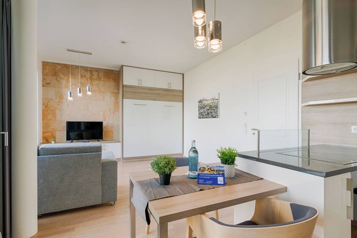 A modern kitchen with a built-in stove, a dining area with a wooden table and chairs, and a grey sofa in the background.