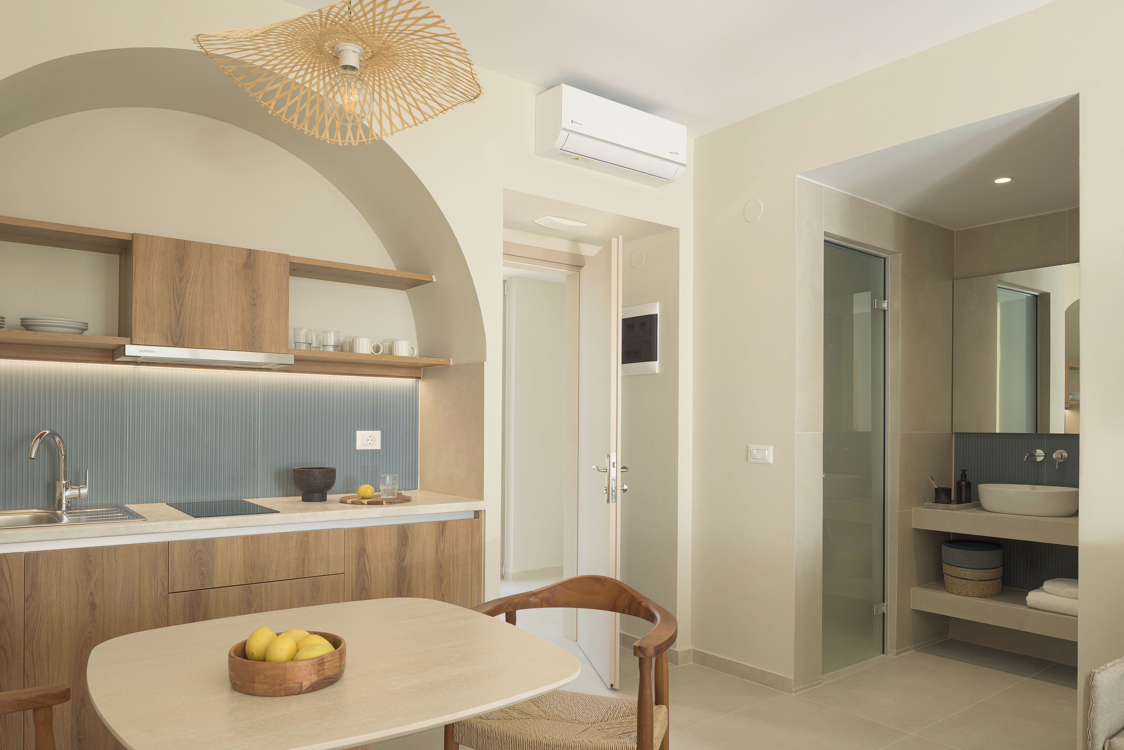 A modern kitchen with a dining area, featuring a wooden table with a bowl of fruit, a built-in sink, and a hanging light fixture.
