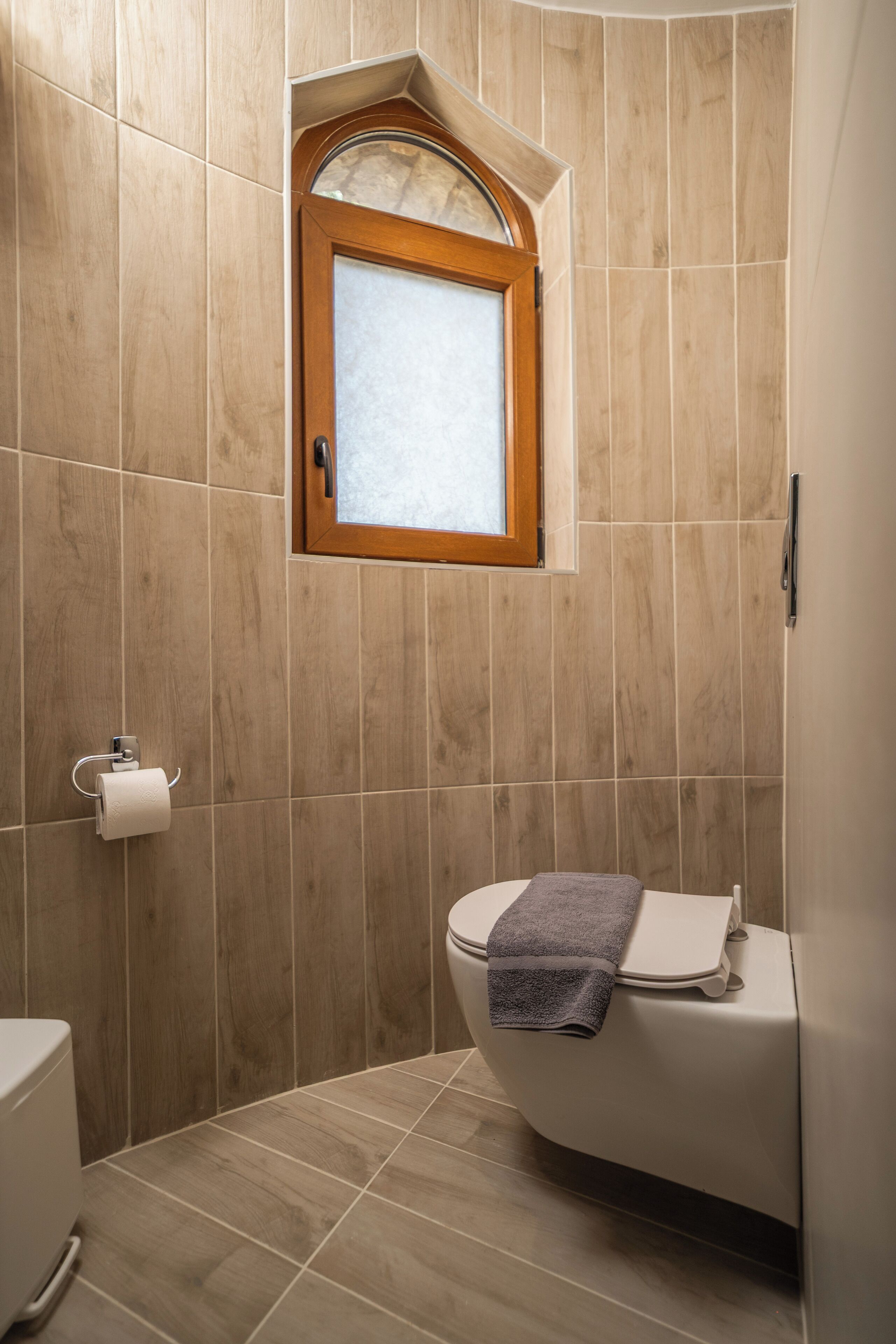 A bathroom with a wooden-framed window, a toilet with a towel, and a toilet paper holder on the wall.