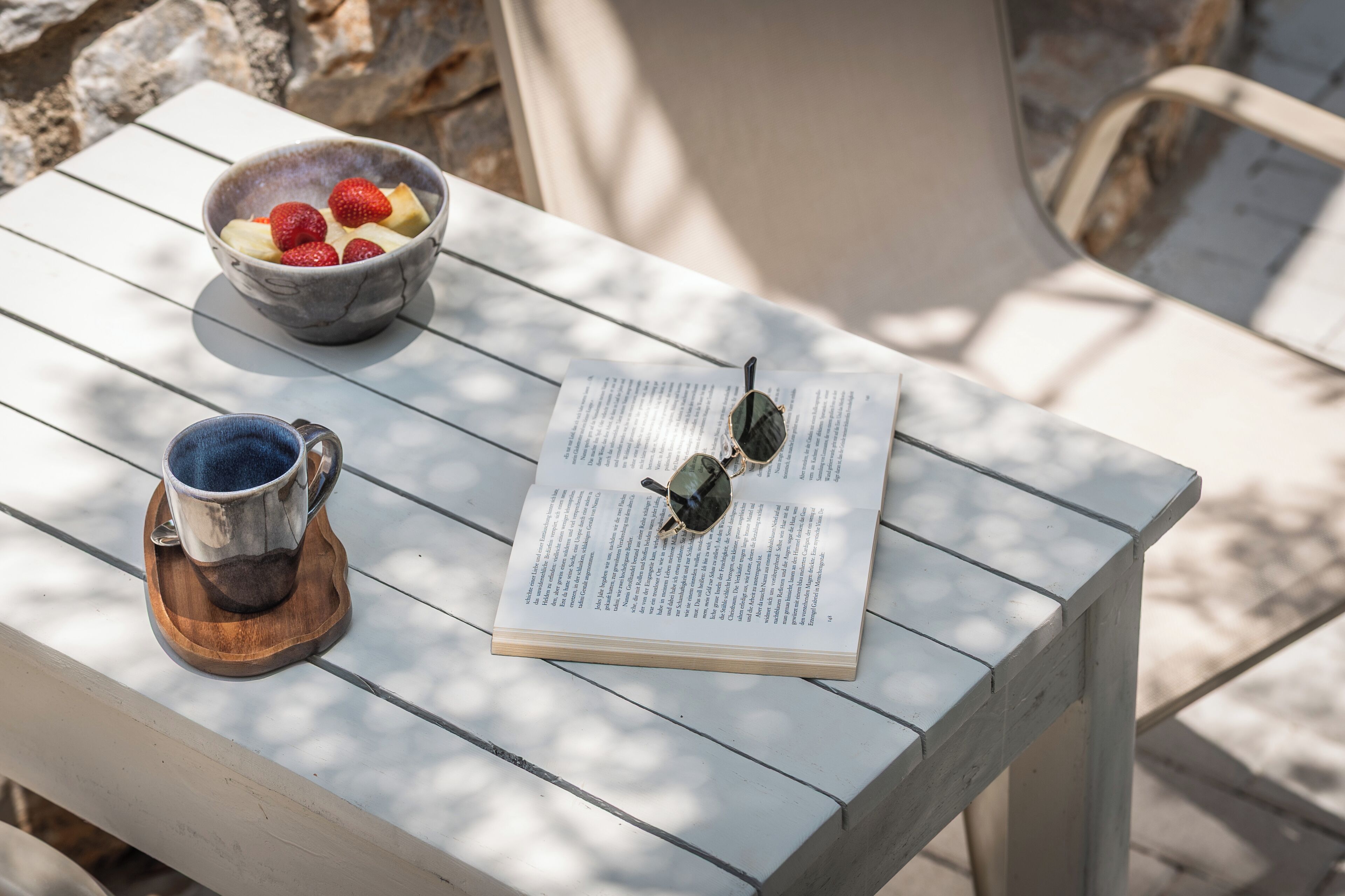 Une table en bois sur laquelle se trouvent un saladier rempli de fruits, une tasse et un livre, avec une paire de lunettes de soleil posée sur le dessus.