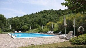 Una piscina con baldosas de piedra, rodeada de sillas de descanso y sombrillas, con árboles y colinas como fondo.
