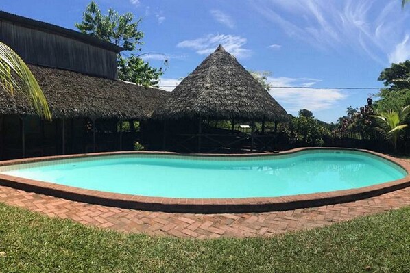 Ein Swimmingpool mit Backsteinrand, umgeben von Gras und strohgedeckten Gebäuden.