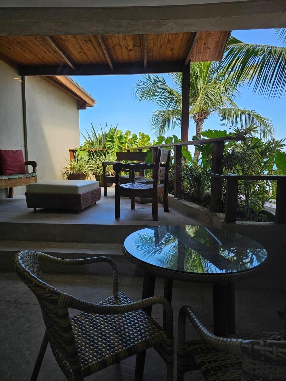 An outdoor patio with wicker furniture, a glass table, and a view of palm trees.