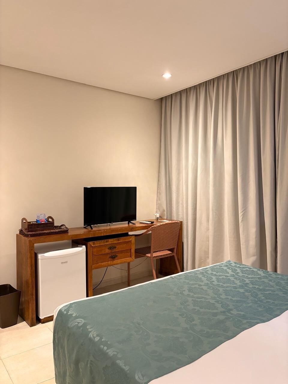 A hotel room with a bed, a desk with a television, a small refrigerator, and a window with curtains.