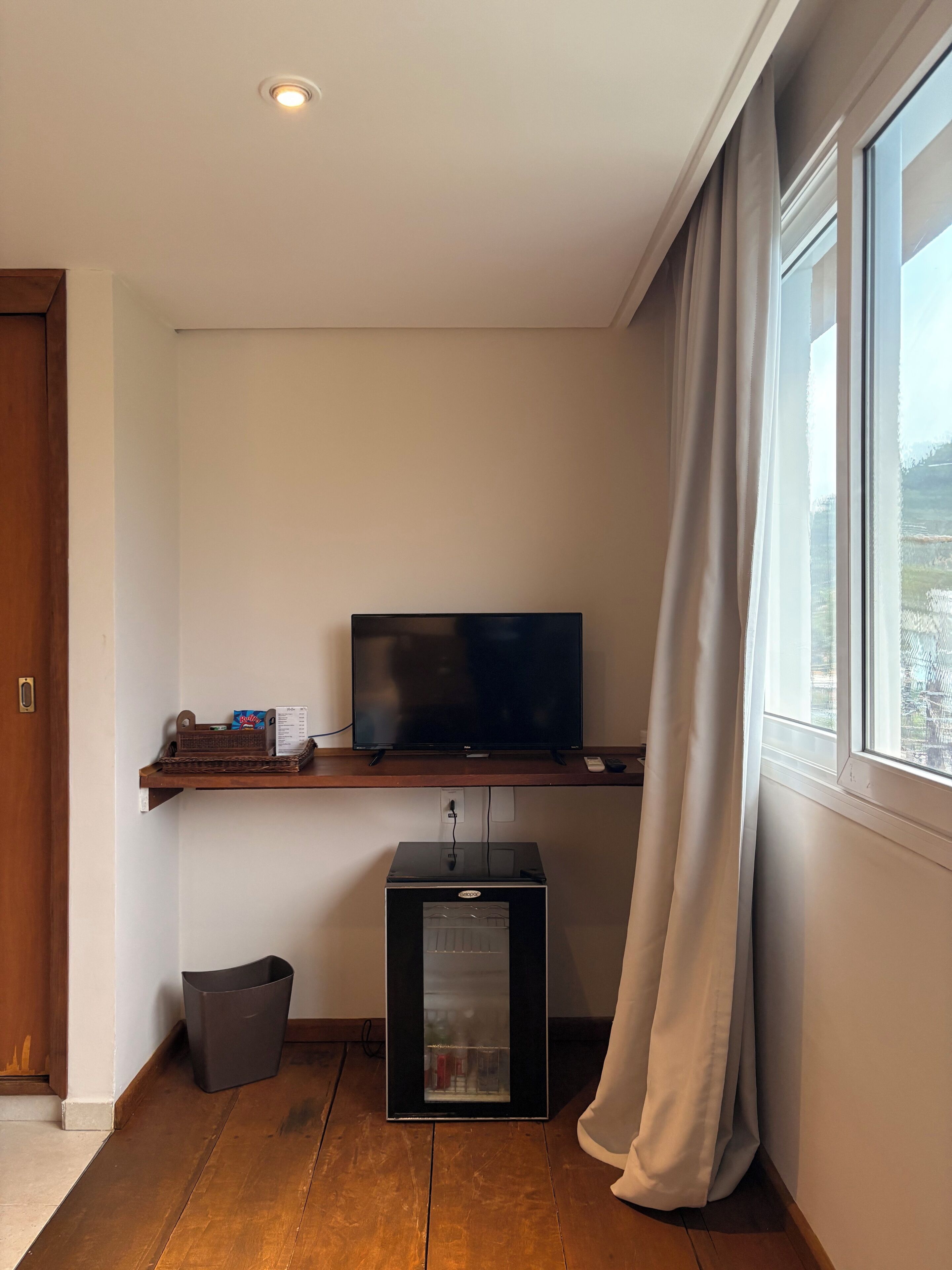 A room with a television on a wooden shelf, a small black cabinet, a waste bin, and a window with curtains.