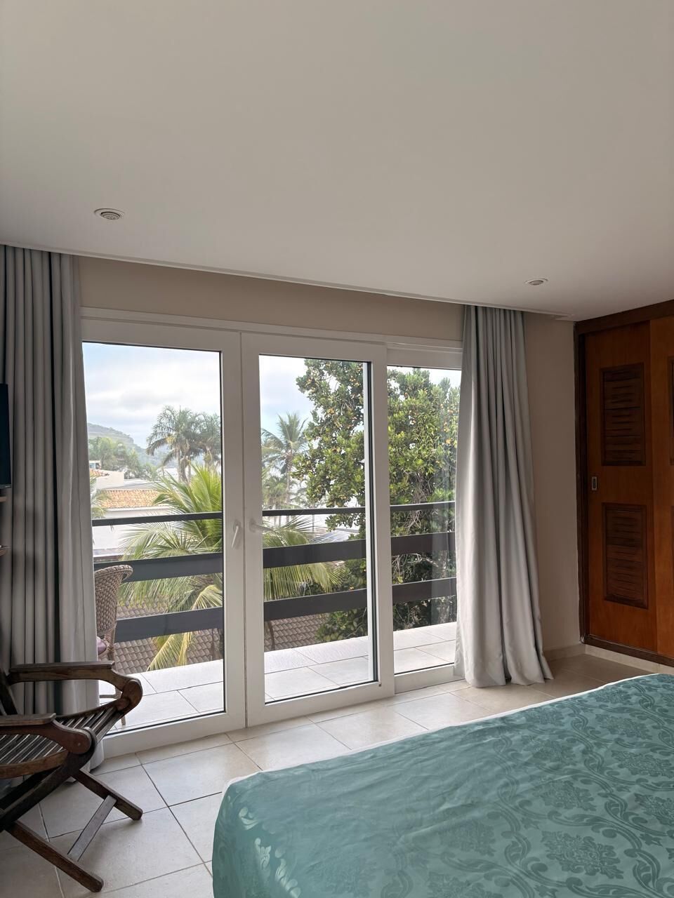 A bedroom with a bed, a chair, a sliding glass door to a balcony, and a view of palm trees and buildings.