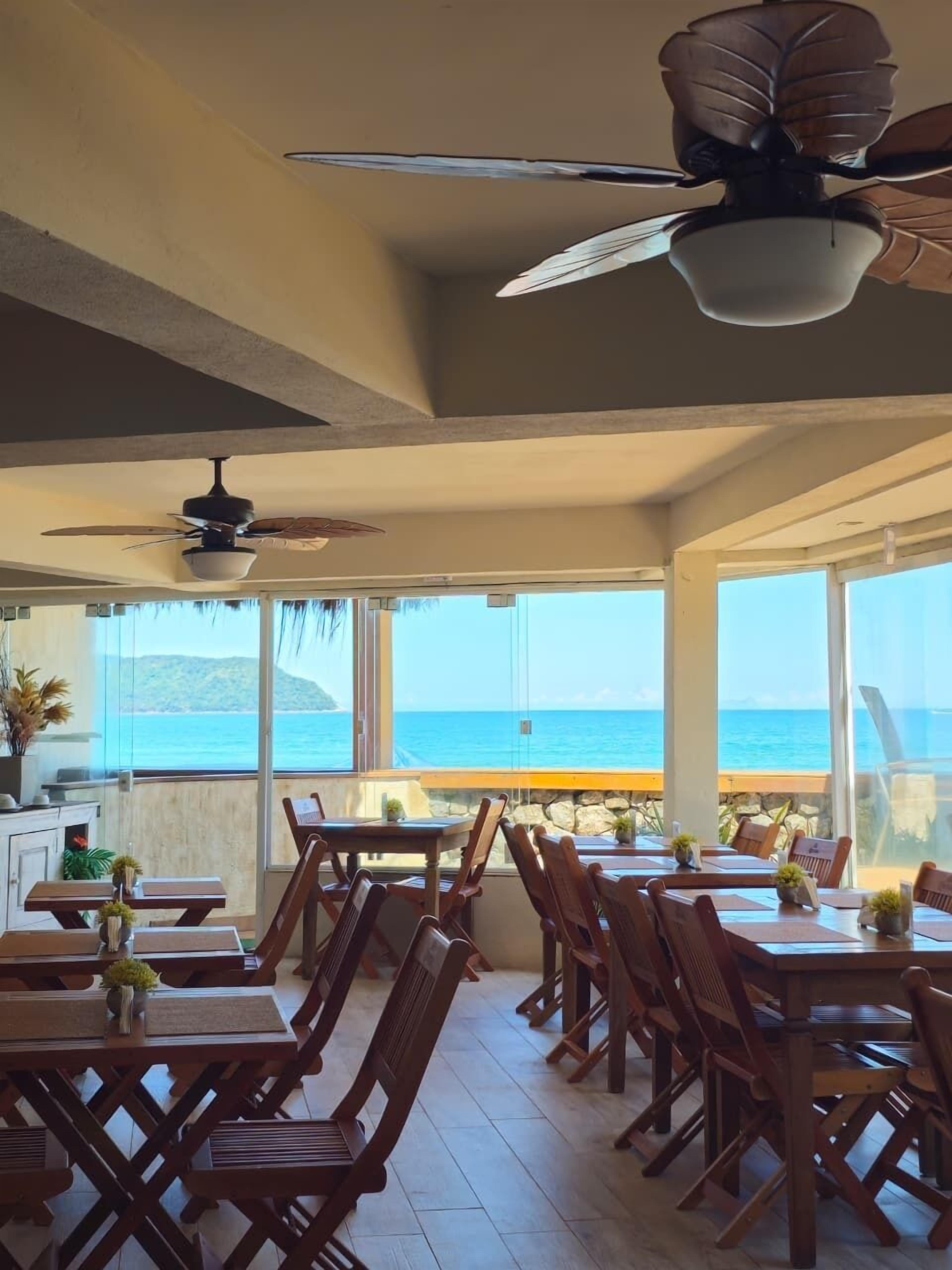 Un restaurante con mesas y sillas de madera, ventiladores de techo y vista al océano.